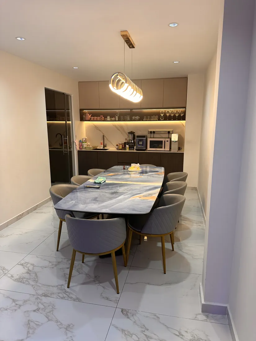 Dining table at Cedar Avenue under a sculptural pendant, with lit display shelving and under-cabinet lighting in the kitchen beyond