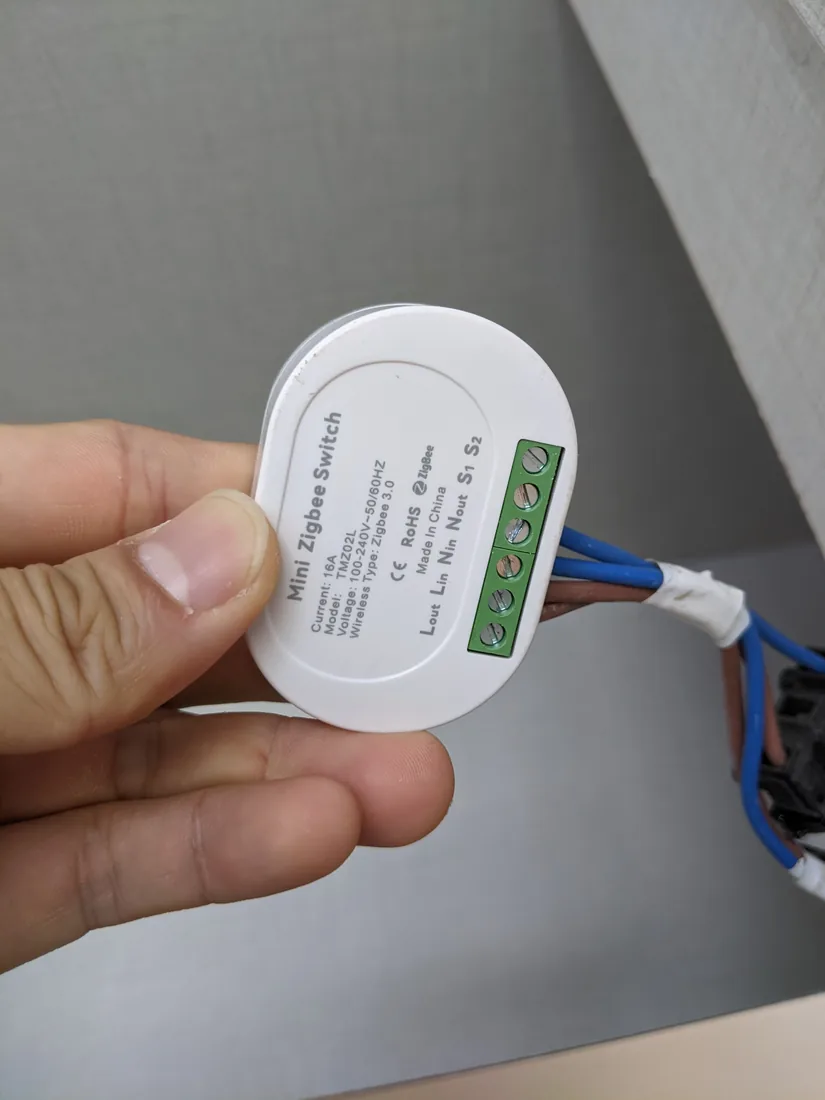 A small round in-wall Zigbee relay module held in one hand, with its wiring tails connected.