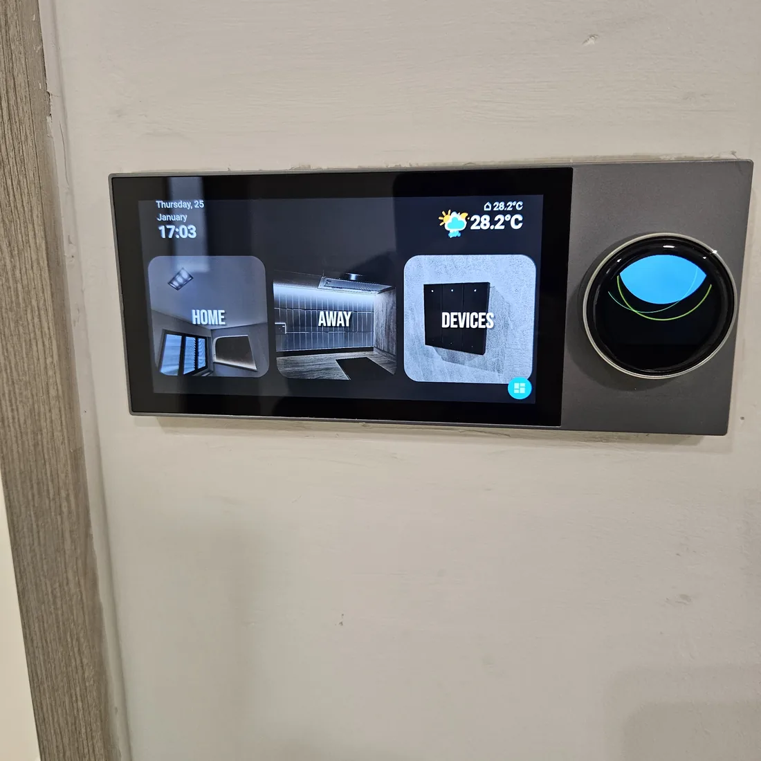 A wall-mounted control panel showing Home, Away and Devices buttons alongside a round dial and a 28.2C room temperature.