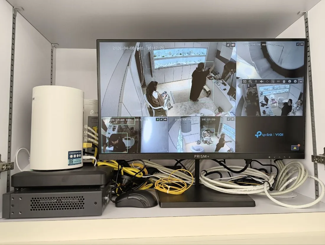 A wall-mounted monitor showing a multi-camera CCTV view above a recorder, network switch and patch cables.